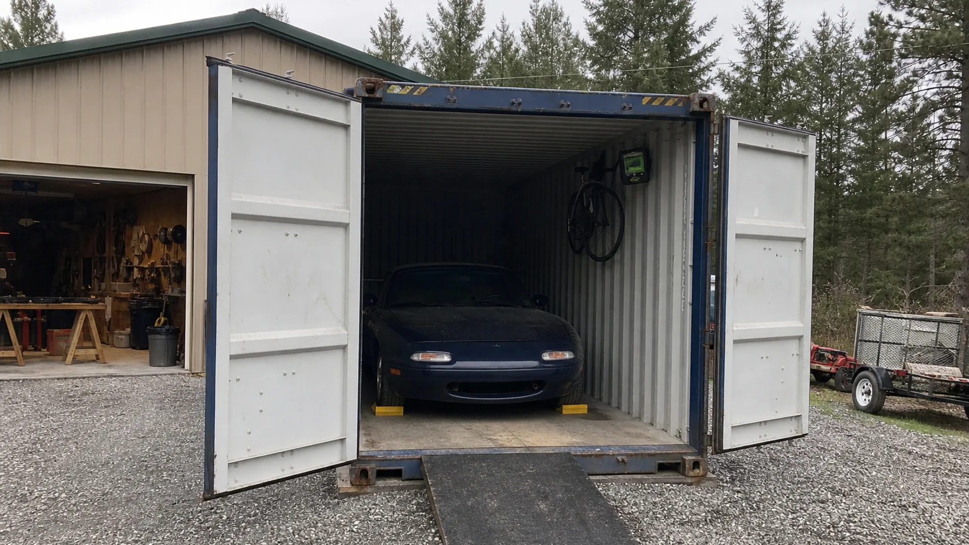Compact roadster stored inside a used 20 foot shipping container beside a rural workshop